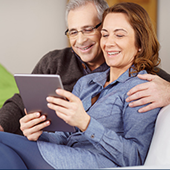 An image of a man and woman smiling while cuddled on a couch and using a tablet.